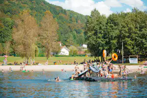 Badesee mit Schwimmfloß und Rutsche, viele Kinder im Wasser, im Hintergrund Bäume und ein kleines Gebäude am Ufer.