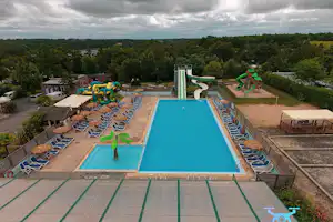 Camping-Pool mit türkisfarbenem Becken, vielen Liegen, Sonnenschirmen und grünen Wasserrutschen, umgeben von Bäumen.