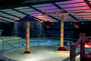 Beheiztes Poolbecken mit Wasserfontänen unter einer Überdachung, im Hintergrund Lampen und Glasgeländer.