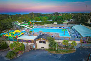 Großer Camping-Pool mit blauen Bahnen und bunten Wasserrutschen, umgeben von Bäumen und mehreren Gebäuden.