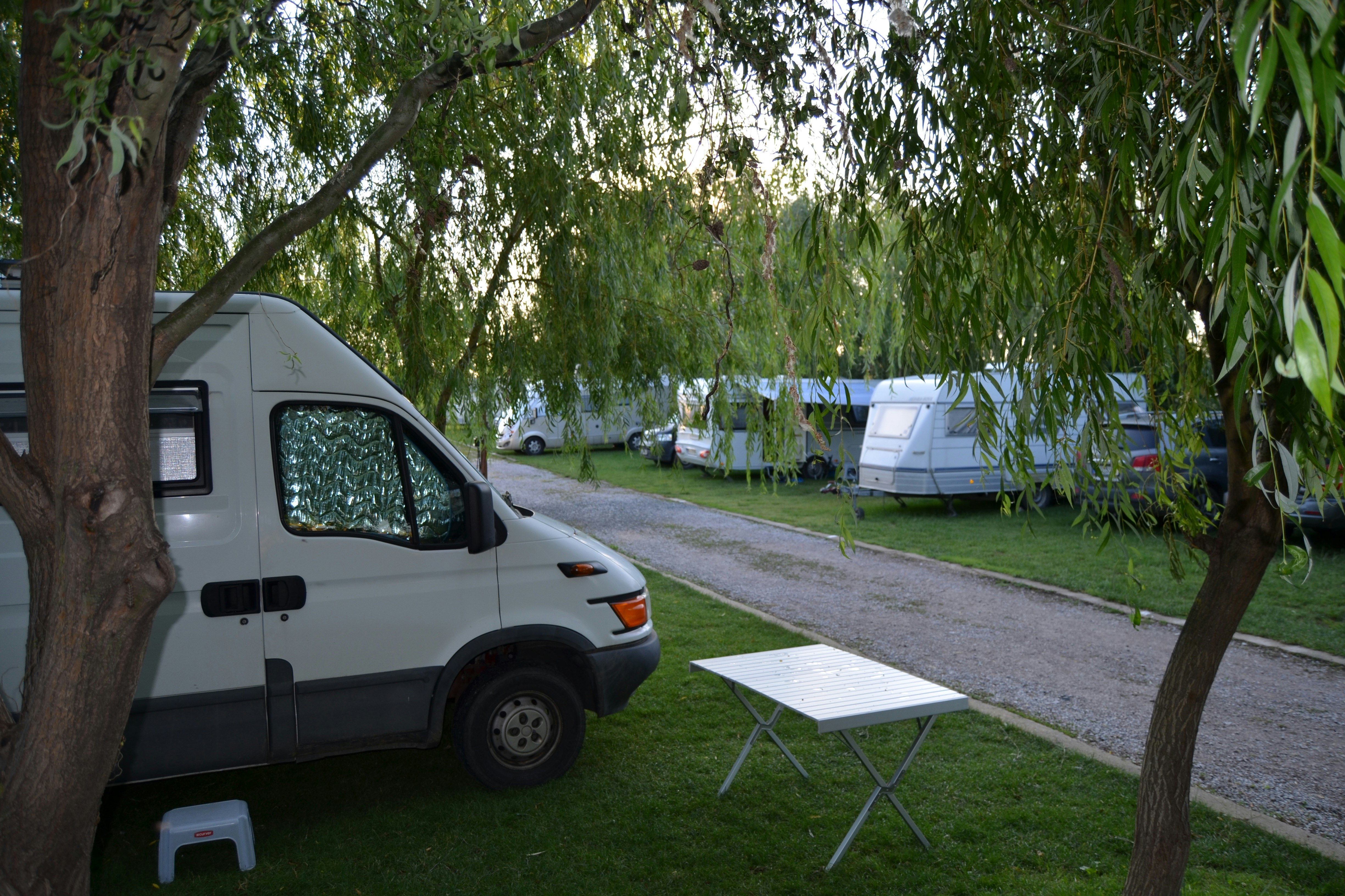 Weißes Wohnmobil am Campingplatz, daneben ein Tisch und Stühle auf dem Stellplatz, im Hintergrund Zelte und Bäume.
