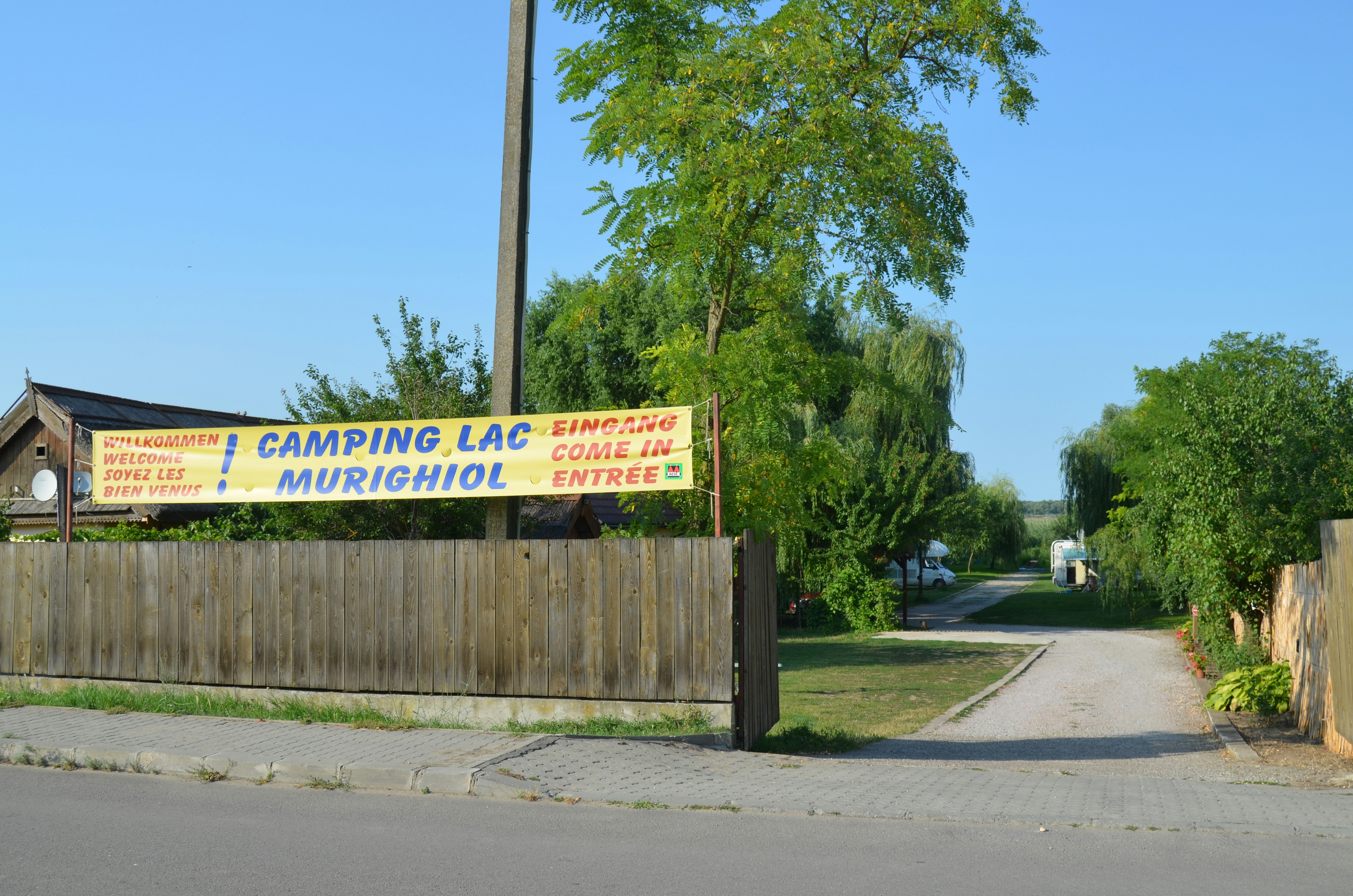 Willkommensbanner am Eingang eines Campingplatzes, dahinter Straße, Holzzaun, Bäume und Wohnwagen im Grünen.
