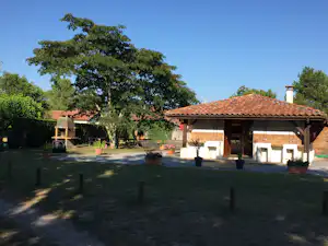 Holzgebäude mit überdachter Terrasse, Sitzgelegenheiten und Tisch auf dem Campingplatz, umgeben von Bäumen und Rasen.