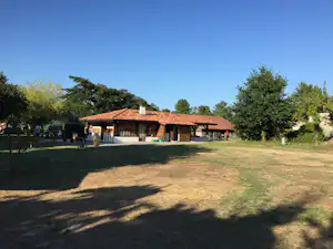 Campinghaus mit rotem Ziegeldach und Terrasse, umgeben von Rasen und Bäumen, im Hintergrund weitere Gebäude.