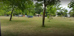 Campingwiese mit Bäumen und mehreren Stellplätzen, im Hintergrund Fahrzeuge und kleine Gebäude.