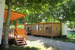 Mobilheim mit Holzveranda und Treppe, umgeben von Bäumen, mit Kiesfläche am Eingang.