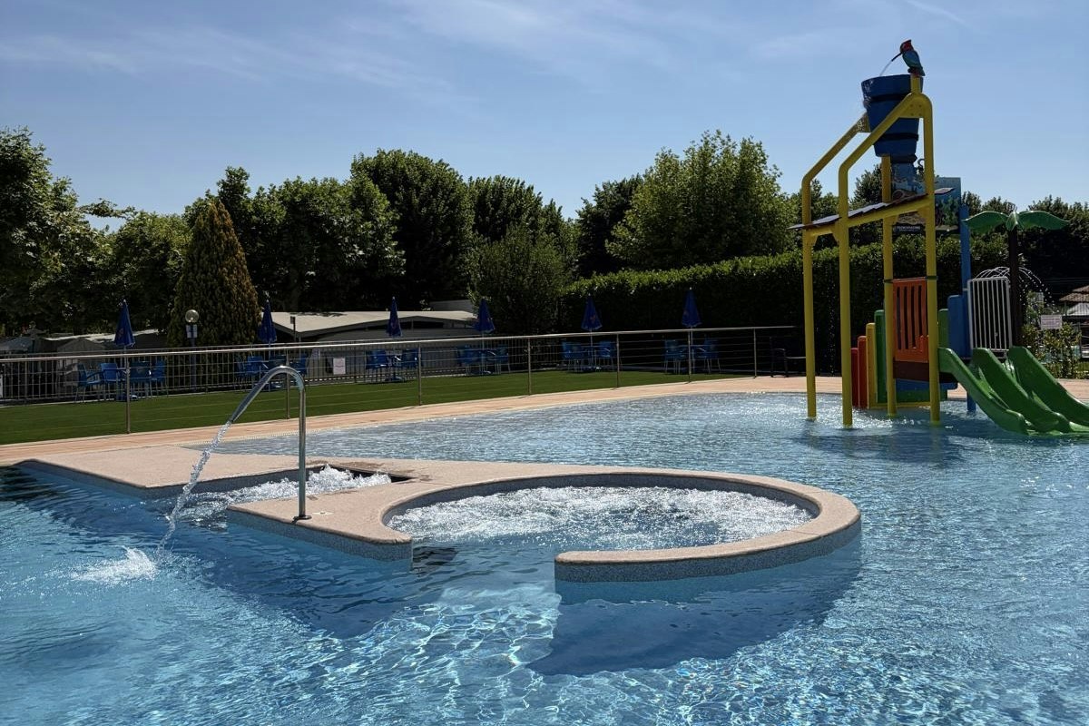 Schwimmbecken mit Rundbecken und Sprudler, gelber Spielturm mit Rutsche, umgeben von Bäumen und blauen Absperrungen.