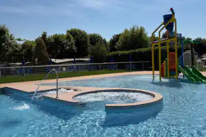 Schwimmbecken mit Rundbecken und Sprudler, gelber Spielturm mit Rutsche, umgeben von Bäumen und blauen Absperrungen.