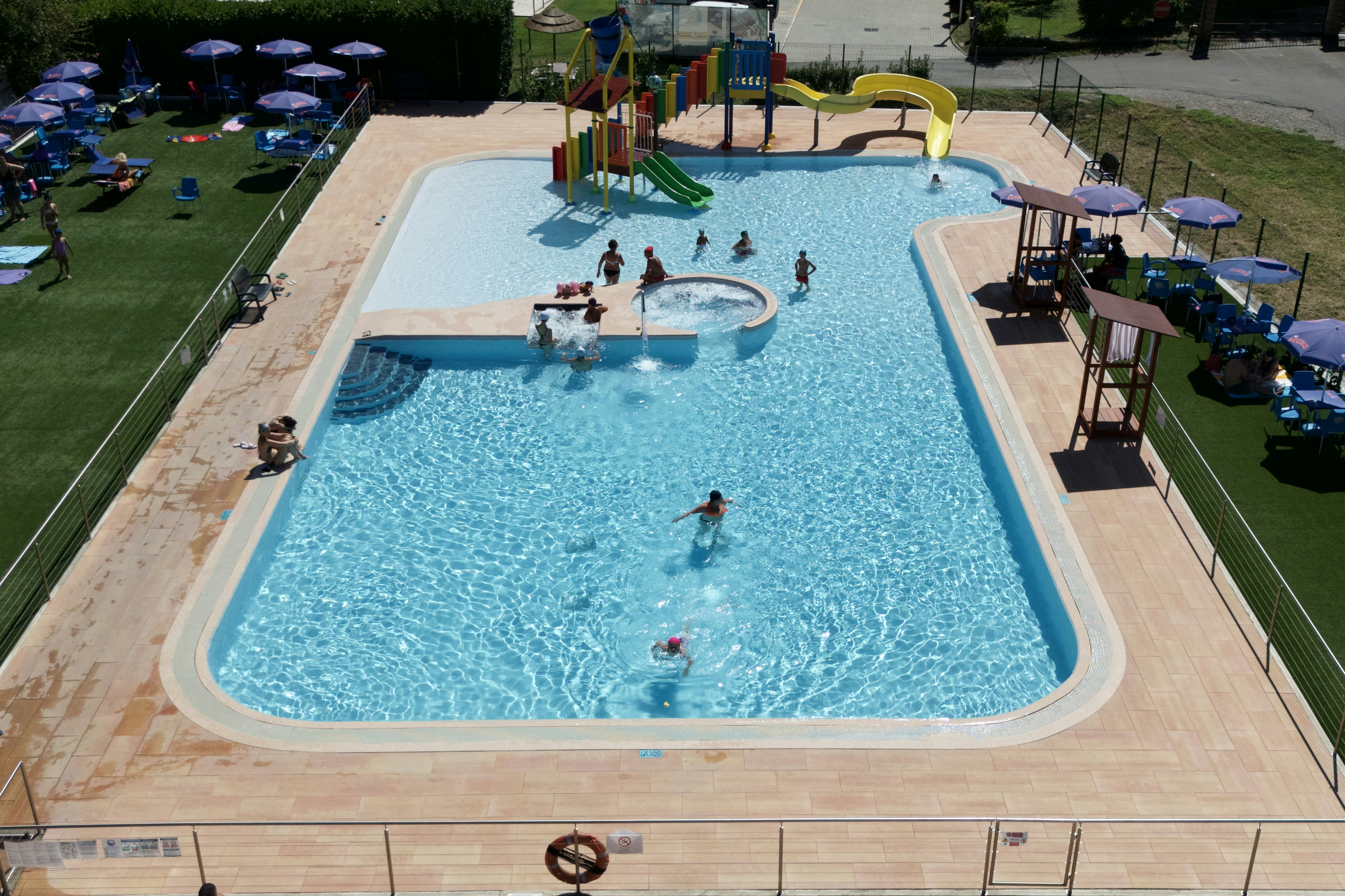 Großes Schwimmbecken mit mehreren Personen, Liegen und Sonnenschirmen auf einer Terrasse, daneben ein Spielbereich im Campingplatz.