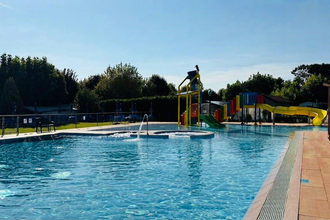 Schwimmbad mit blauem Wasser, Edelstahlgeländer und Rutsche, daneben Liegen und Spielplatz mit bunten Kletterelementen.
