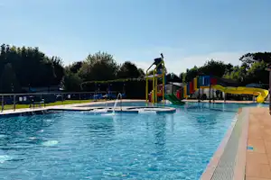 Schwimmbad mit blauem Wasser, Edelstahlgeländer und Rutsche, daneben Liegen und Spielplatz mit bunten Kletterelementen.