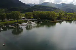 Ruhiger See mit Stegen und Booten, Campingplatz am Ufer, umgeben von Bäumen und Bergen im Hintergrund.