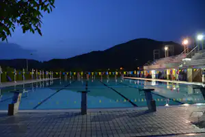 Beleuchtetes Schwimmbecken auf dem Campingplatz am Abend, mit Startblöcken, Liegen und Zäunen, im Hintergrund ein bewaldeter Hügel.