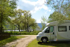 Wohnmobil auf einem Campingplatz am Weg, umgeben von Bäumen und Wiese, im Hintergrund Berge und Himmel.