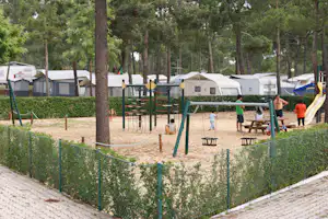 Campingplatz mit Wohnwagen und Zelten, Spielplatz mit Schaukeln und Klettergerüst, Hecken und gepflasterten Wegen zwischen den Stellplätzen.