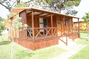 Holzbungalow mit Veranda, Eingangstür und Fensterfront, umgeben von Rasen, Bäumen und Sträuchern im Campingbereich.