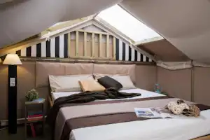 Bett in einem Zelt mit gestreifter Holzverkleidung, Kissen und Decke, daneben Nachttischlampe und Wasserflasche.