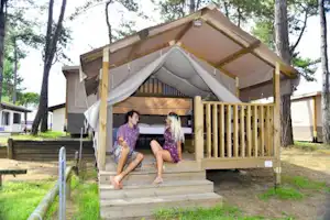 Holzpodest mit Zeltunterkunft und Vorhang, umgeben von Bäumen; daneben Sitzbank und Zaun am Campingplatz.