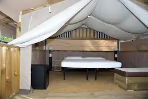 Zelt mit weißen Planen, zwei Betten und Holzkisten auf einem Campingplatz-Stellplatz, daneben ein schwarzer Schrank.