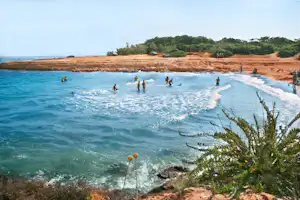 Badegäste im Meer mit Surfbrett, Blick auf den Sandstrand, Dünen und Autos am Ufer.