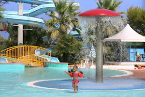 Wasserspiel im Pool mit Spritzfontäne, roter Rutsche, Palmen und Liegen am Beckenrand.