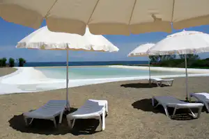 Pool am Campingplatz mit Sonnenschirmen, weißen Liegen und Blick auf das Meer hinter dem Zaun.