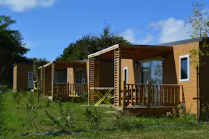 Holz-Bungalow am Campingplatz mit Veranda, Stufen und Geländer, umgeben von Rasen und Bäumen.