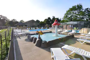 Beheizter Pool mit Springbrunnen, Liegen und Sonnenschirmen auf dem Campingplatz, im Hintergrund Bungalows und Bäume.