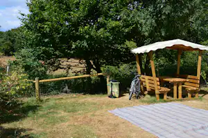 Holz-Pavillon mit Sonnendach und Sitzbank auf einem Campingplatz, daneben Wäschekorb und Rasenfläche mit Bäumen im Hintergrund.