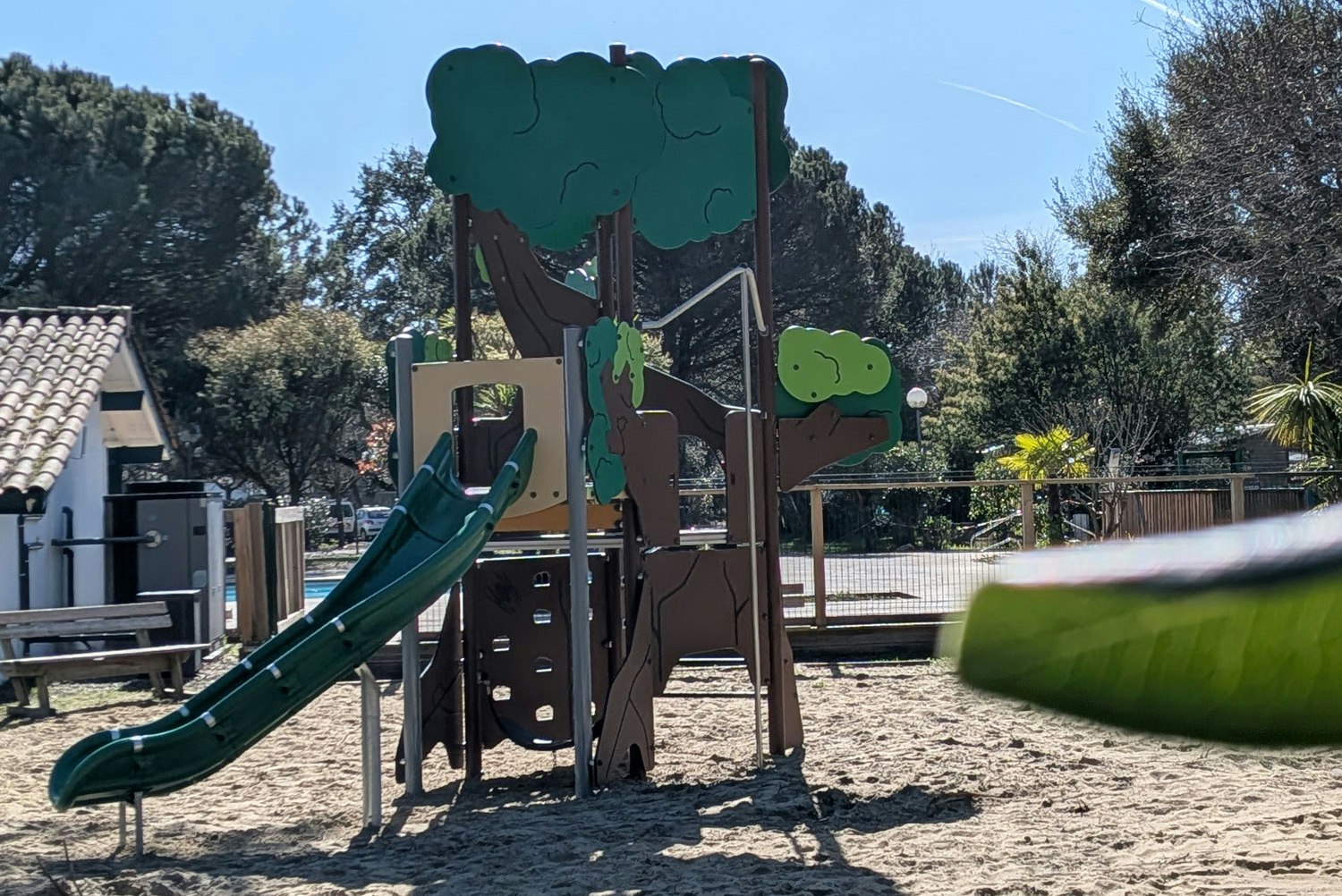 Spielplatz mit Rutsche und Kletterturm aus Holz auf dem Campingplatz, im Hintergrund Bäume und ein Zaun.