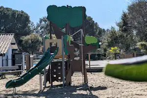 Spielplatz mit Rutsche und Kletterturm aus Holz auf dem Campingplatz, im Hintergrund Bäume und ein Zaun.