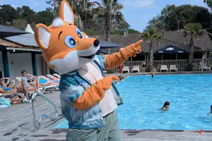 Bunte Camping-Maskottchenfigur steht am Holzdeck neben dem Pool, im Hintergrund Liegen und Gebäude mit Palmen.