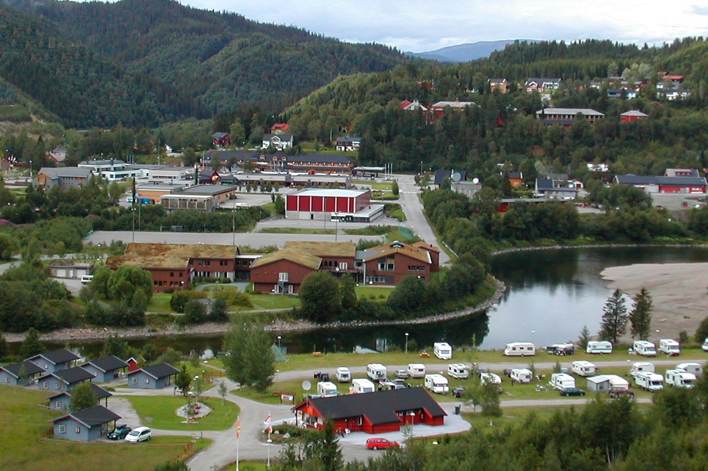Campingplatz mit Wohnwagen und Zelten am Fluss, Blick auf Wege, Gebäude und grüne Hügel im Tal.