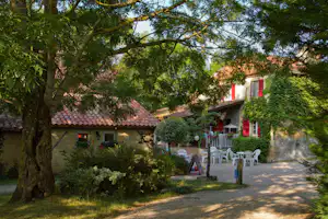 Haus mit roter Fensterfront und Terrasse, weiße Gartenstühle und Tisch im Schatten von Bäumen und Hecken.