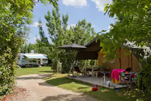Campingzelt mit überdachtem Sitzbereich, daneben Tisch und Stühle, Rasen und Hecken, im Hintergrund Wohnmobil auf dem Stellplatz.
