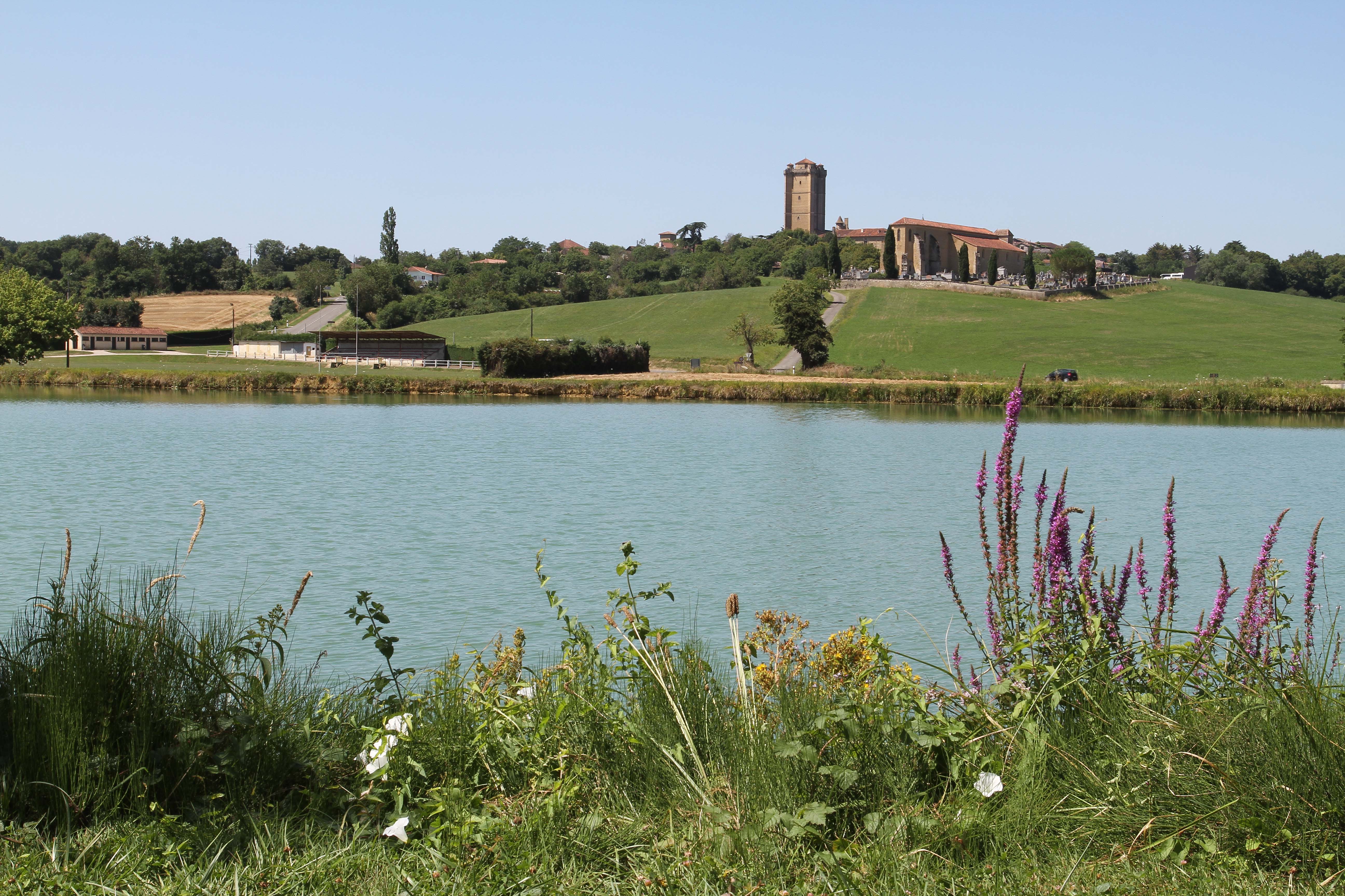 Ruhiger See am Campingplatz mit grünen Hügeln, Ufergras und einem Gebäude mit Turm in der Ferne.