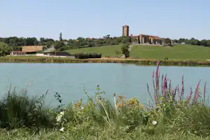 Ruhiger See am Campingplatz mit grünen Hügeln, Ufergras und einem Gebäude mit Turm in der Ferne.