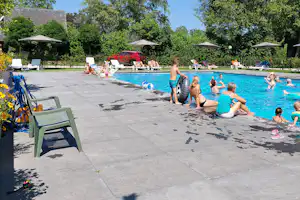 Kinder und Erwachsene im Pool eines Campingplatzes, mit Liegen und Sonnenschirmen am Beckenrand, im Hintergrund Bungalows und Bäume.