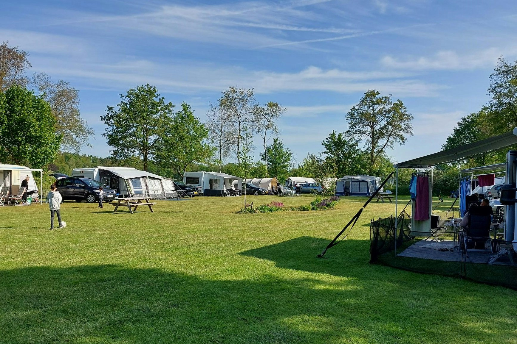 Campingplatz mit Rasenfläche, mehreren Wohnwagen und Zelten, Sitzgelegenheiten und Sonnenschirmen entlang eines Weges.