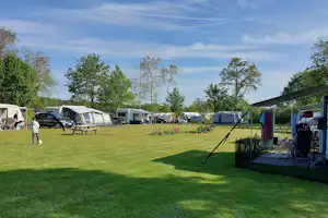 Campingplatz mit Rasenfläche, mehreren Wohnwagen und Zelten, Sitzgelegenheiten und Sonnenschirmen entlang eines Weges.