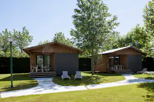 Zwei Holz-Campingunterkünfte mit Terrasse, Sitzgelegenheiten und Rasenfläche, umgeben von Bäumen und einem Weg.