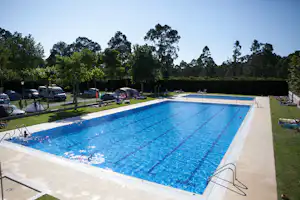 Großer Camping-Pool mit blauem Wasser, Liegen am Rand und geparkten Autos neben dem Becken, umgeben von Bäumen.