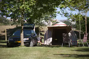 Campingzelt mit Markise und Stühlen auf dem Rasen, daneben ein Auto unter Bäumen.