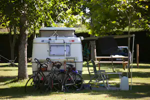 Wohnmobil mit Fahrrädern am Heck, Campingstühle und Tisch im Schatten der Bäume auf einer Wiese.