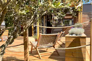 Holzterrasse mit zwei Gartenstühlen und Holzkiste mit Pflanzen, im Hintergrund ein Olivenbaum und ein Gebäude.