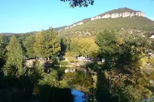 Campingplatz am Fluss mit Bäumen, Stellplätzen und Wohnwagen, im Hintergrund ein bewaldeter Hügel mit Felswand.