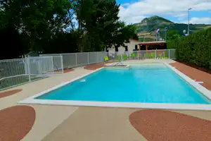 Schwimmbecken auf einem Campingplatz mit Liegen, Zaun und Bäumen; im Hintergrund ein Berg und ein klarer Himmel.