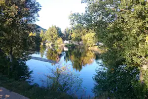 Blick auf den Fluss mit Spiegelung, Bäume am Ufer und geparkte Wohnmobile auf dem Campingplatz.