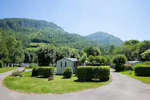 Kleines Wohngebäude auf einem Campingplatz mit Hecken, Kieswegen und Rasenfläche vor grünen Hügeln.