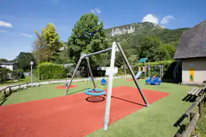 Spielplatz am Campingplatz mit rotem Fallschutzboden, Schaukel und Spielgeräten, Hecken und Blick auf Berge.